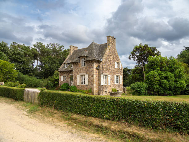 Plevenon, France - July 20, 2024: Charming traditional stone breton house with a sloped roof, surrounded by greenery and a well-maintained hedge.