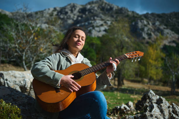 A woman enjoys playing her guitar outdoors, surrounded by a peaceful mountain landscape. Her expression reflects serenity and connection with nature, capturing a moment of musical tranquility.