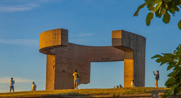 Gijon, Spain. 26th August 2024. A group of people are standing in front of a large, curved structure. The sky is blue and there are a few clouds. Scene is peaceful and serene, as the people are enjoying the view and taking pictures