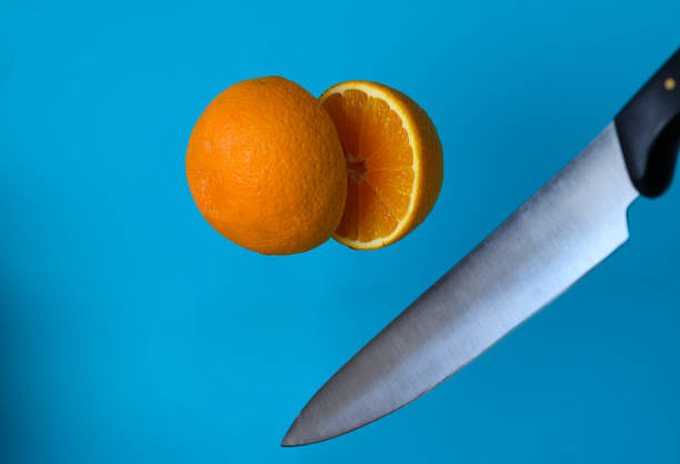 A sharp knife hovers above a whole orange, with a sliced section revealing its juicy interior on a bold blue surface.
