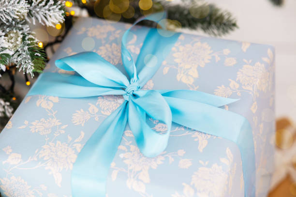 A carefully wrapped gift featuring floral-print wrapping paper and a delicate blue ribbon sits under a beautifully decorated Christmas tree adorned with lights and ornaments.
