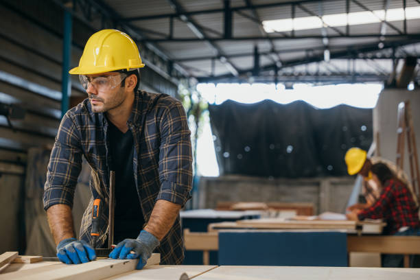 istock Construction worker in a yellow hard hat and blue gloves cutting wood on a workbench. Ideal for woodworking, carpentry, and construction projects, showcasing tool use and craftsmanship 2183387223