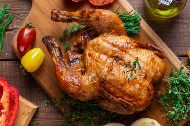 istock A perfectly roasted whole chicken garnished with herbs on a wooden board, accompanied by sliced lemons, red and yellow bell peppers, grilled tomatoes, fresh rosemary, and olive oil. 2183401297