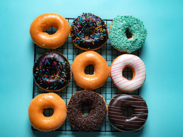 istock A grid of assorted donuts is displayed on a black cooling rack against a bright turquoise background. The donuts include glazed, chocolate, sprinkle-topped, and coconut-covered varieties. 2183401336