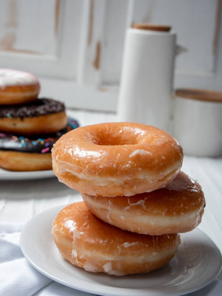 istock stack of glazed donuts placed on a white plate. In the background, there is another plate with a variety of donuts, including one with chocolate sprinkles. The scene is set in a light, airy kitchen. 2183401664
