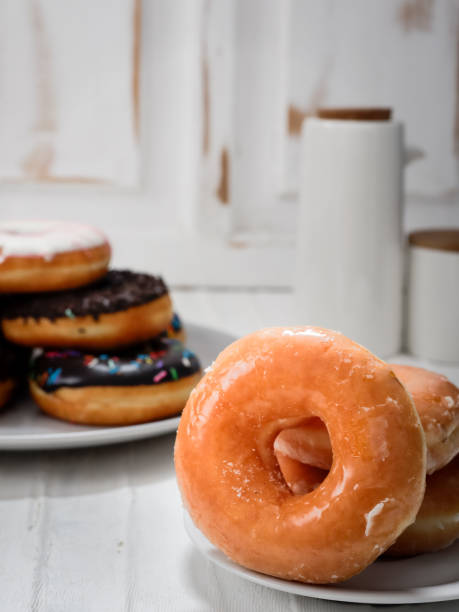 istock stack of glazed donuts placed on a white plate. In the background, there is another plate with a variety of donuts, including one with chocolate sprinkles. The scene is set in a light, airy kitchen. 2183401665