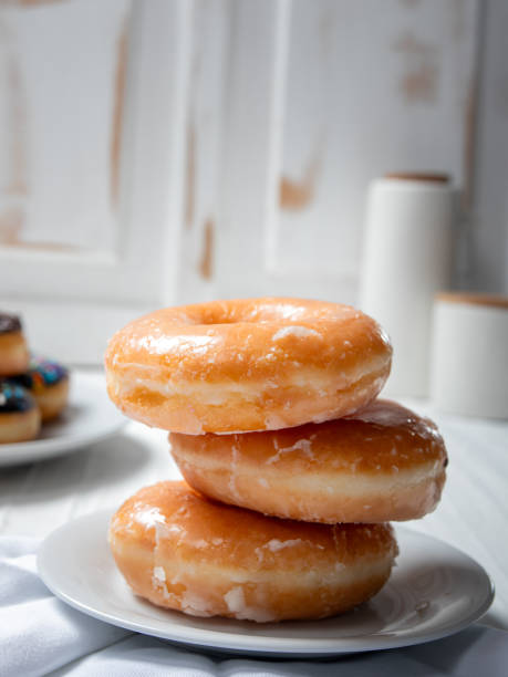 istock stack of glazed donuts placed on a white plate. In the background, there is another plate with a variety of donuts, including one with chocolate sprinkles. The scene is set in a light, airy kitchen. 2183401693