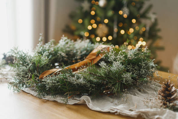 istock Making stylish Christmas rustic wreath. Wreath with evergreen branches, ribbon, scissors on rustic table on background of atmospheric christmas tree. Winter holiday preparations 2183402634