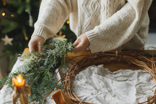 istock Hands holding evergreen branches, making stylish christmas rustic wreath on table with pine cones, twine, scissors and candle on background of christmas tree. Winter holiday preparations 2183402884