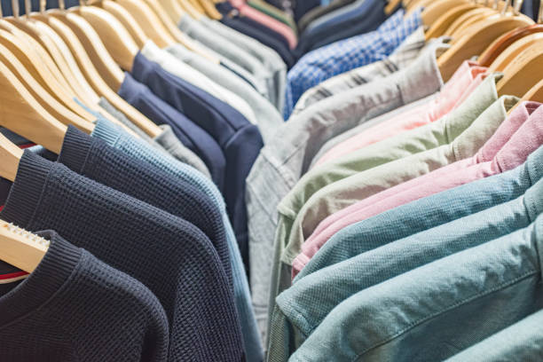 Menswear Array: A Spectrum of Men's Fashion The image presents a neatly organized selection of men's clothing on wooden hangers, showcasing a spectrum of colors and styles. The garments range from casual to more formal attire, with a palette that includes shades of blue, gray, and a spectrum of greens and pinks. The orderly arrangement and the variety of colors and textures suggest a well-curated collection designed to appeal to a diverse male clientele. The focus on color and orderliness emphasizes the store's commitment to presentation and the shopping experience. This photograph captures the essence of a men's clothing section in a retail store, inviting customers to explore the array of fashion options available. colorful check shirts on sale at a clothing store stock pictures, royalty-free photos & images