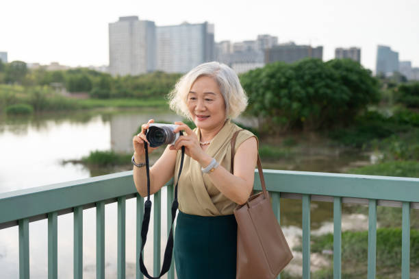 An older Asian woman wearing beige and green stands on a bridge taking photos with a camera, enjoying the serene outdoor environment under a cloudy sky.
