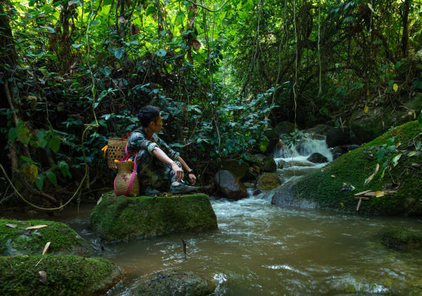 A man wearing camouflage uniform and carrying a traditional rattan backpack sits beside a jungle stream, quietly admiring the flowing water, reflecting the harmonious coexistence of man and nature