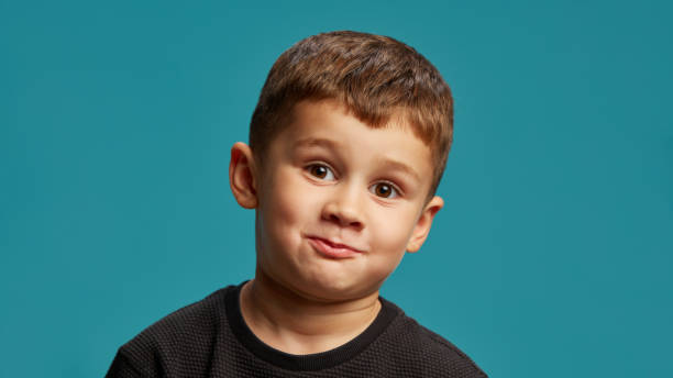 istock Little boy pulls playful face, smirk with his lips puckered, giving comical and mischievous vibe against blue studio background. 2183458784