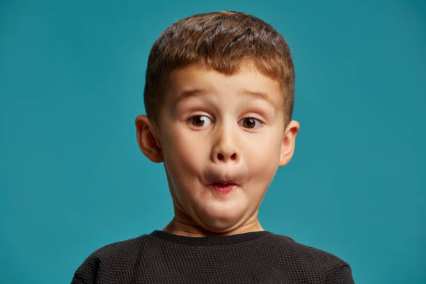 istock Boy makes comical expression with puffed cheeks, his eyes wide open in surprise against blue studio background. Childhood curiosity. 2183458815
