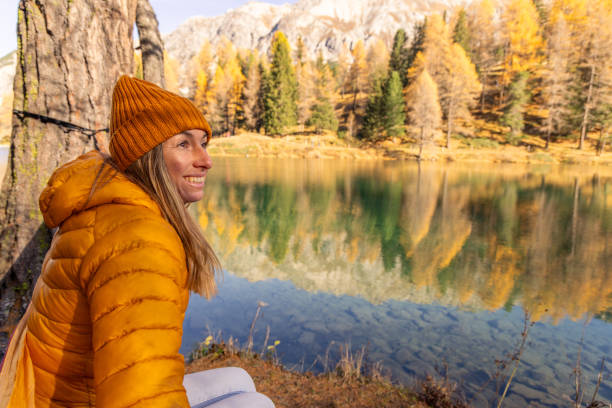 Woman in a vibrant orange jacket enjoys a peaceful moment in a hammock, surrounded by autumn colors and reflecting lake waters against a mountain backdrop.