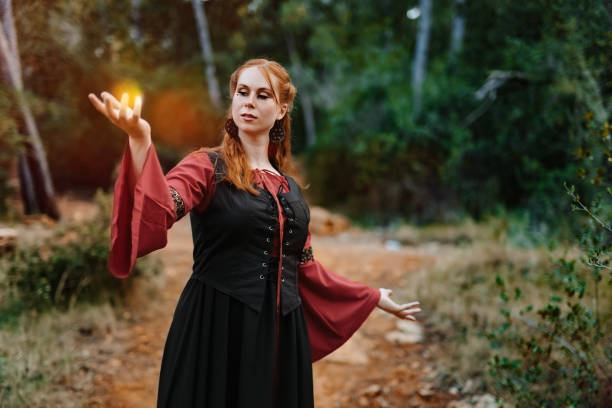 istock Beautiful red-haired medieval fantasy witch in the deep  forest playing with a glowing golden light 2183479655