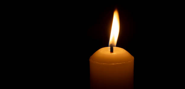 istock A single burning candle flame or light glowing on a big white candle isolated on black or dark background on table in church for Christmas, funeral or memorial service 2183489368
