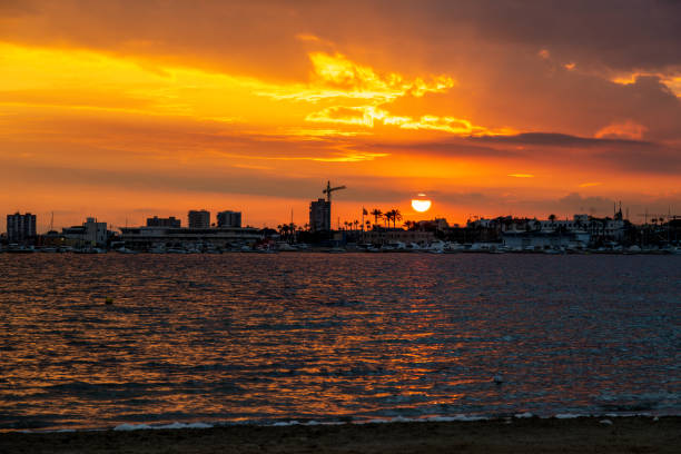 The setting sun lights up the horizon in vibrant orange and yellow hues, with dramatic clouds and silhouettes by the water.
