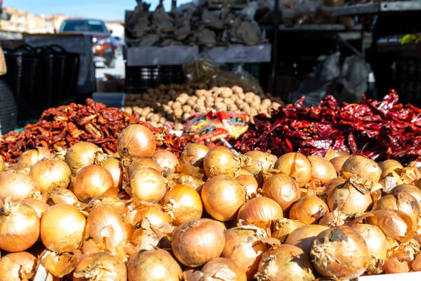 A market stand features large quantities of onions, dried peppers, and walnuts in the background, set outdoors.
