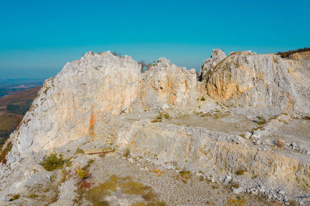 Aerial view of a rugged limestone quarry with exposed rocky terrain and distinct geological formations under a clear blue sky. The barren land and rough stone textures highlight the area's mining activity and natural erosion.