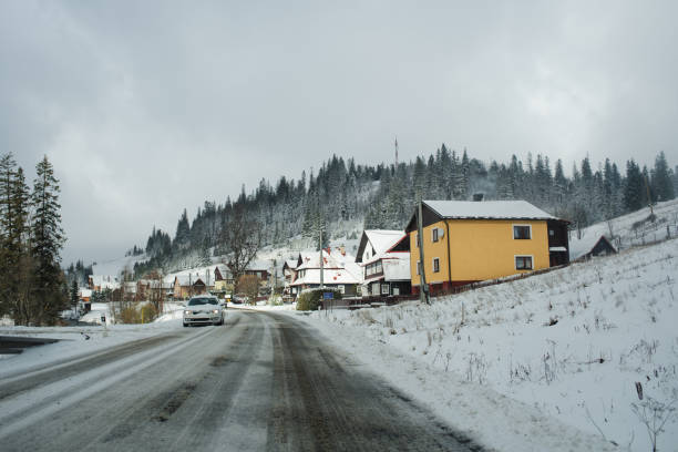 A car drives down a snowy road through a quiet village nestled at the base of a forested mountain. Snow-covered houses and cloudy skies add to the serene winter atmosphere.