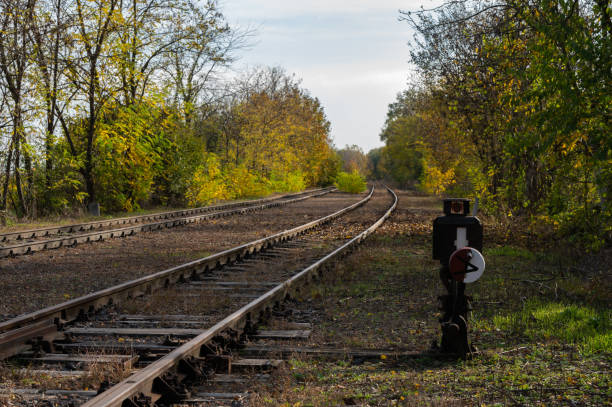 An autumn landscape featuring railroad tracks leading into the distance, flanked by trees with fall foliage. A point indicator stands beside the tracks, marking the pathway in this peaceful, natural setting.