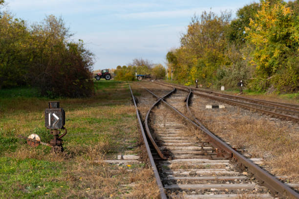 A rural railroad scene in autumn, showing tracks with a point indicator in the foreground. A tractor and hay bales are visible in the distance, blending agricultural and railway elements in a tranquil countryside setting.