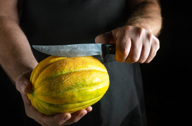 In a dimly lit kitchen, a skilled chef holds a bright melon, poised with a sharp knife, ready to create a delicious dish as shadows dance around them