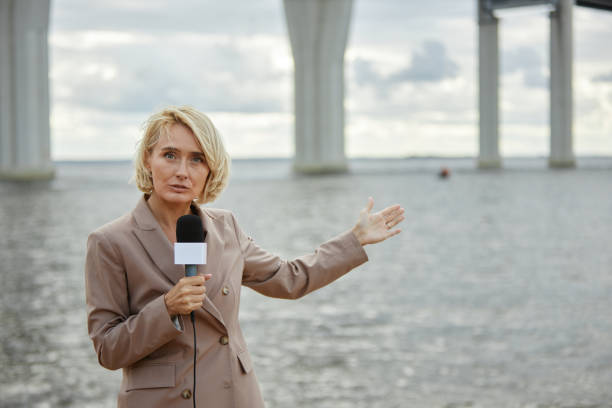 Waist up portrait of young blonde woman as news reporter speaking to microphone and gesturing to vast water landscape in background, copy space
