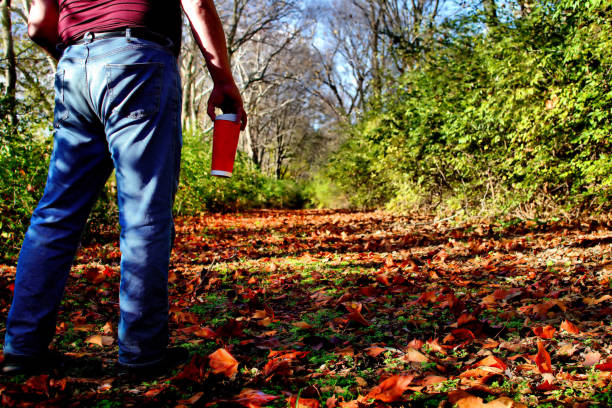 Man in blue jeans carrying a red coffee cup stands at the start of a leaf covered path that leads through the woods in autumn.