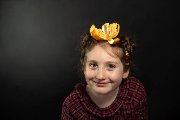 Young girl in red plaid dress against dark background creatively balances orange slices on her head. Her hair styled in two buns complements the playful aesthetic. Vibrant orange fruit contrasts dramatically with the dark backdrop, showcasing her cheerful expression and whimsical approach.