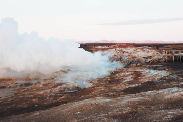 This stunning photograph captures the ethereal Gunnuhver hot springs in Iceland at sunset. Clouds of steam rise dramatically from the ground as geothermal energy forces hot air through the earth's crust, creating a mystical atmosphere. The landscape is painted in autumnal shades of deep orange and red, blending beautifully with the warm hues of the setting sun. The sky mirrors the landscape with brilliant reds and oranges, casting a soft, fiery glow over the scene. This captivating moment highlights the raw, vibrant energy of Iceland’s geothermal landscape, set against the enchanting colors of a fall sunset.