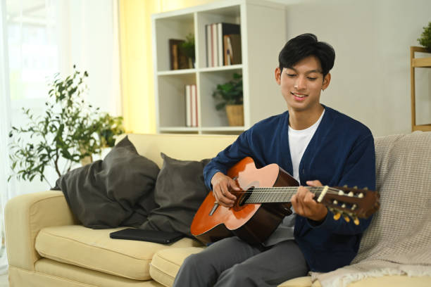 istock Smiling young adult man tuning and practicing acoustic guitar on sofa 2183625636