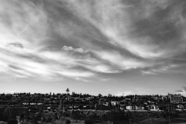A captivating black-and-white view of Kigali's hillside residences beneath dramatic, swirling clouds. This image showcases the contrast between the built environment and the expansive sky, capturing the quiet elegance of Kigali's architecture and natural surroundings. The monochrome effect emphasizes the texture and depth of the clouds, adding a timeless quality to the Rwandan cityscape.