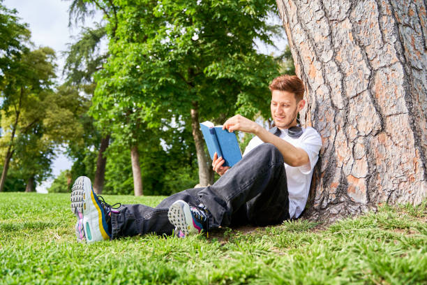 istock A man sits on the grass in a park, leaning against a tree while absorbed in reading a book, enjoying a tranquil moment in nature. 2183635452