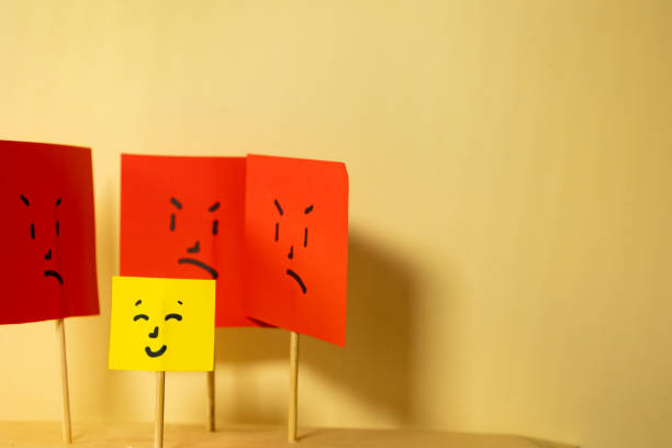 Horizontal image of yellow paper with smiling square face surrounded by angry square faces, concept of social rejection and discrimination with copy space