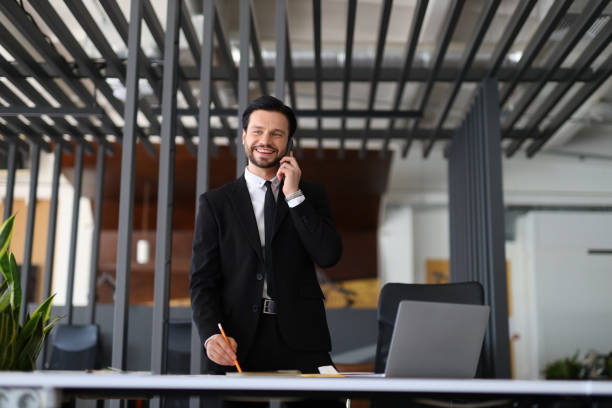 istock A man in a suit is talking on a cell phone while standing in front of a desk. He is smiling and he is in a good mood 2183649427
