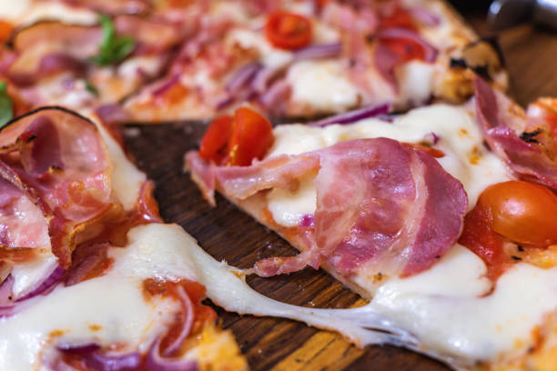 A slice of prosciutto pizza is served, showcasing melted mozzarella, fresh tomatoes, and aromatic basil, inviting customers in a lively dining area.