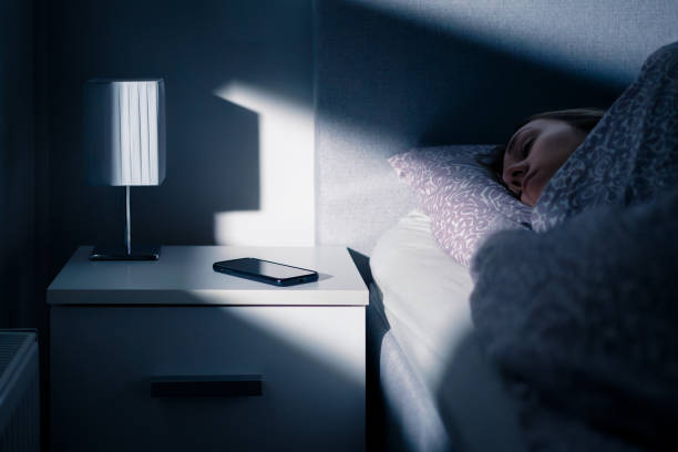 Phone next to bed while sleeping. Mobile on table and nightstand at night. Woman and shut down cellphone at bedside. Light from window. Young person asleep in bedroom.