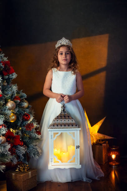 A young girl dressed as a princess holds a glowing lantern next to a beautifully decorated Christmas tree in a cozy setting.