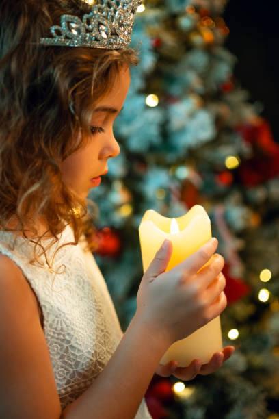 A girl wearing a tiara holds a glowing candle with both hands, standing near a decorated Christmas tree in soft lighting.