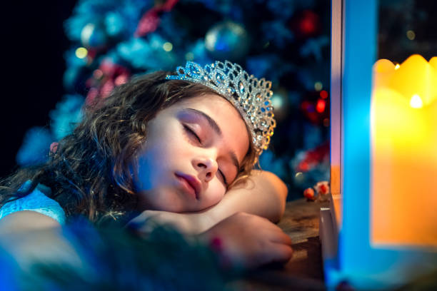 A young girl wearing a tiara sleeps by beautifully decorated Christmas tree.