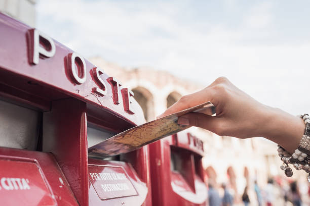 A young woman is seen posting a letter into a red mailbox, with a historic building in the background.