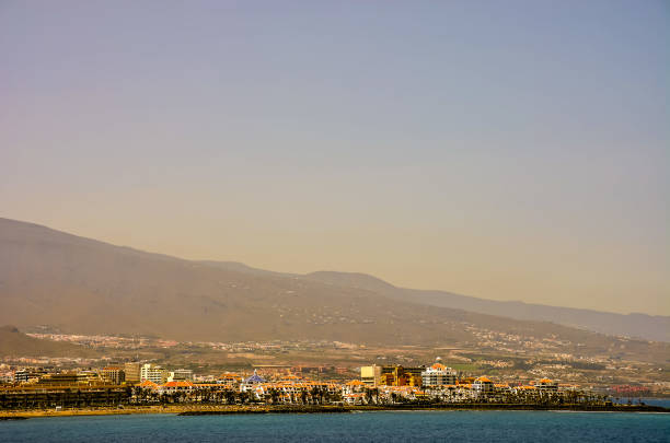 A city with a mountain in the background. The sky is blue and the sun is setting. The city is full of buildings and the water is calm