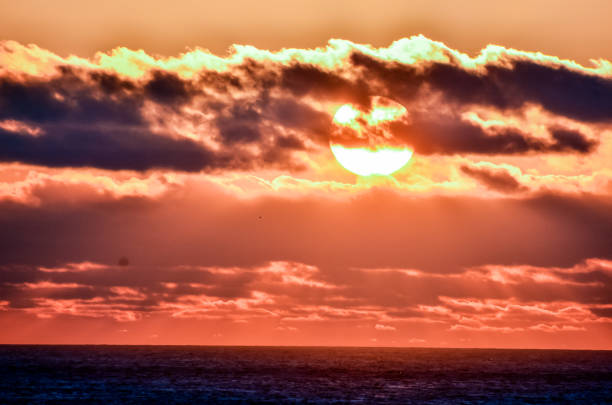 The sun is setting over the ocean, casting a warm glow over the water. The sky is filled with clouds, creating a serene and peaceful atmosphere