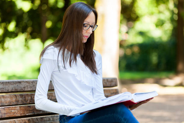 istock Young woman reading a book on park bench 2183723066