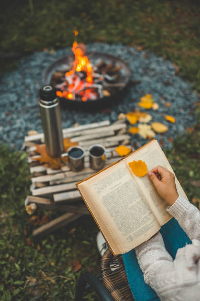 istock A tranquil autumn scene with a person reading a book by a campfire, holding a leaf as a bookmark. A thermos and metal cups rest nearby, enhancing the cozy outdoor atmosphere 2183730942