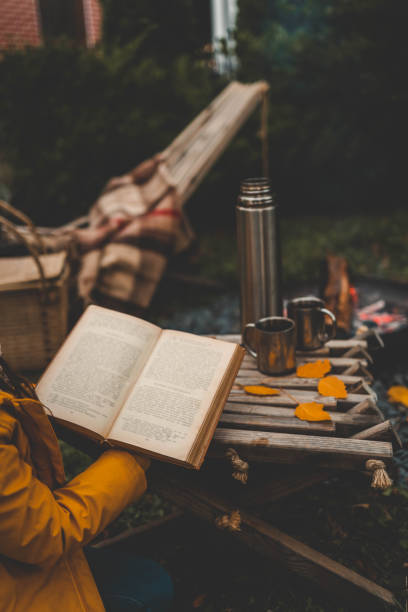 istock A tranquil autumn scene with a person reading a book by a campfire, holding a leaf as a bookmark. A thermos and metal cups rest nearby, enhancing the cozy outdoor atmosphere 2183730959
