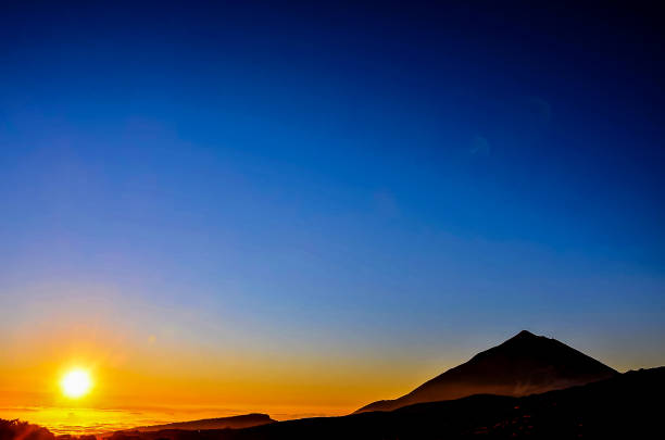 A mountain is silhouetted against a blue sky with a sun setting in the background. The sky is clear and the sun is setting, creating a beautiful and serene atmosphere