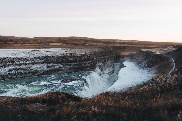 The iconic Gullfoss waterfall in Iceland flows through a partially frozen landscape, capturing the transition between autumn and winter. The surrounding terrain is tinged with warm orange hues, while snow and ice cling to the rocks and cliffs around the cascading waters. Under a pastel-hued sunset, the sky glows with soft purples and oranges, adding a serene, almost ethereal quality to this powerful natural wonder.